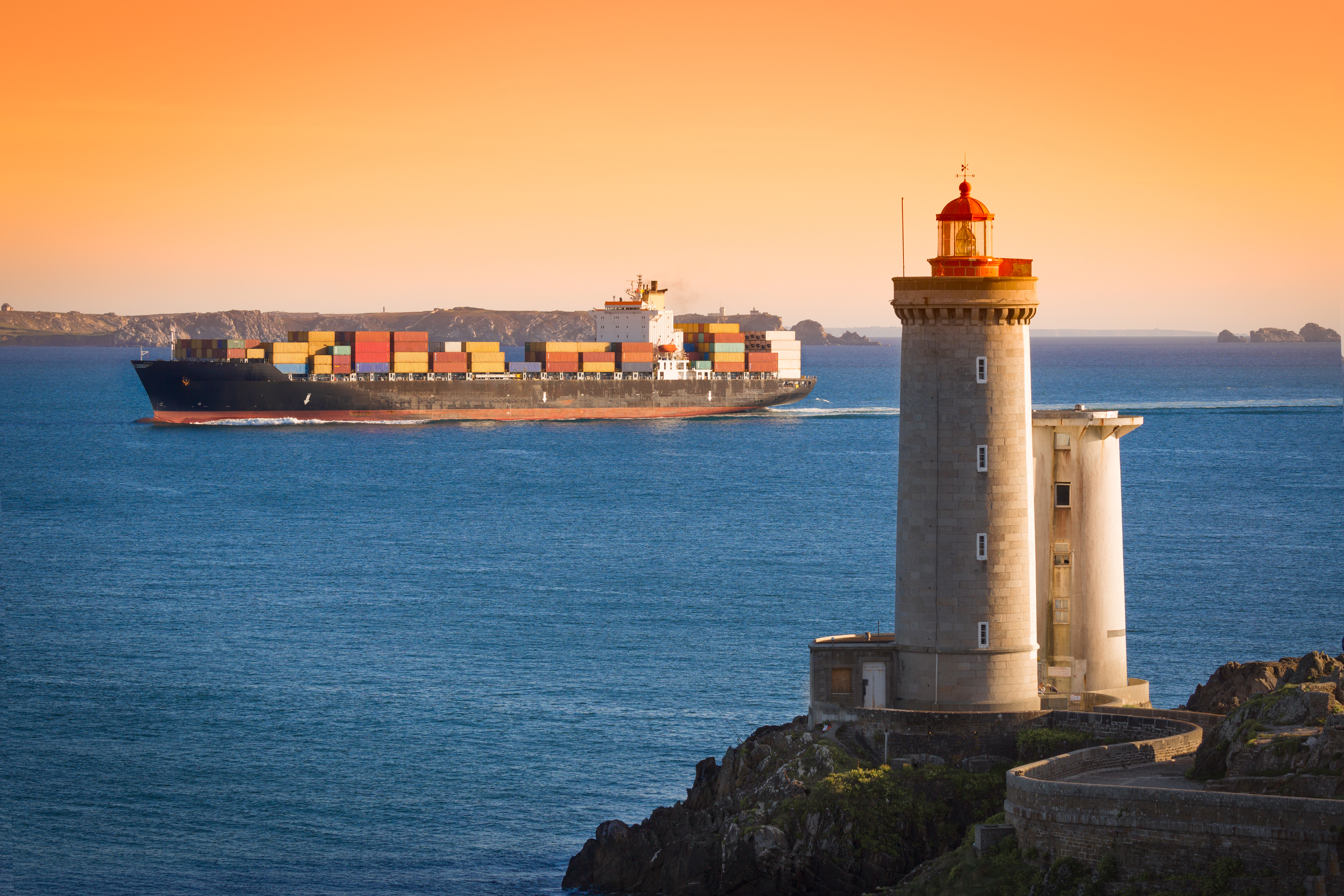 Image of a cargo ship with a lighthouse at sunset in the background.